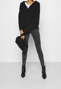 Suéter negro de canalé con un cuello con cremallera, combinado con jeans ajustados gris oscuro y botas negras hasta el tobillo, sosteniendo un bolso negro.