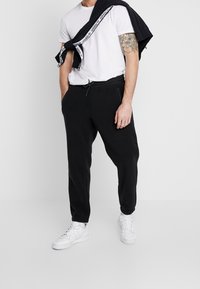 Černé jogger kalhoty s elastickými manžetami a nastavitelným tkaničkovým pasem, spárované s bílým tričkem a černou mikinou přehozenou přes rameno.