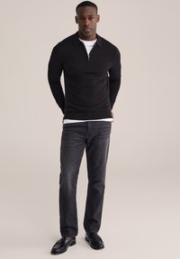 Črn polo pulover z zadrgo, nošen čez belo srajco, kombiniran s temnimi kavbojkami in črnimi loaferji. Preprost dizajn, prilegajoč kroj, minimalna tekstura.