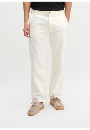 Mann trägt weiße gerade geschnittene Hose, beige Schnürschuhe und schwarzes Hemd, die Hände in den Taschen, vor weißem Hintergrund.