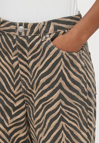 Bikses ar zebras rakstu, kurās izmantots bēšs un melns dizains, teksturēts audums, priekšējās kabatas un pogas aizdare jostasvietā.