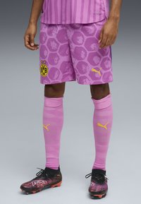 Fotbollsdräkt med rosa tröja med vertikala ränder, lila shorts med hexagonalt mönster, Puma-logotyp och svarta fotbollsskor med färgglada accenter.