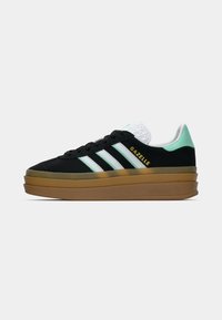 Zapatilla Adidas Gazelle negra con rayas blancas, talón verde menta, logotipo dorado y suela marrón gruesa y estratificada.