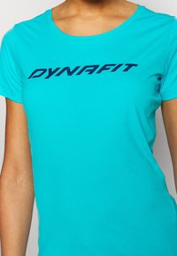 Una camiseta atlética turquesa hecha de tela suave, con la palabra "DYNAFIT" en azul oscuro en el pecho.