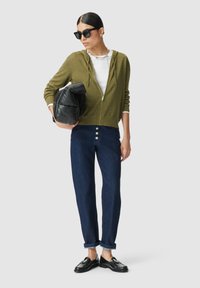 Frau trägt schwarze Sonnenbrille, olivgrüne Kapuzenjacke mit Reißverschluss, weißes T-Shirt, dunkelblaue Jeans mit hochgerollten Säumen, schwarze Loafer und hält eine schwarze Tasche.