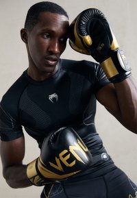 Homme en tenue de sport noire avec des gants de boxe noirs et dorés, posé dans une attitude défensive de boxe sur un fond neutre.