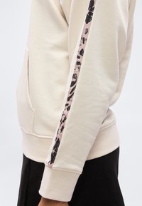 Μανίκι μπεζ φούτερ με λεπτομέρεια ρίγας σε ροζ και μαύρο animal print, φορεμένο με μαύρο παντελόνι.