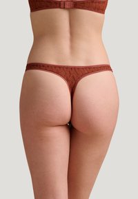 String brun en dentelle avec un motif géométrique, des bretelles étroites et une texture lisse. Comprend un matériel ajustable à la taille.