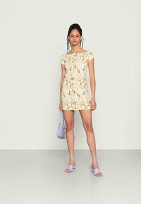 Vestido amarelo curto com mangas curtas, apresentando um padrão espiral em tons de roxo e castanho. Combinado com acessórios em lavanda, incluindo uma bolsa e sandálias.