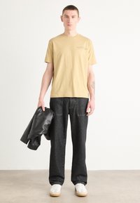 Beige bomulls t-shirt med logotyp, tillsammans med mörk denim byxor med vida ben. Svart läderjacka hålls i vänster hand, vita sneakers med beige sulor.