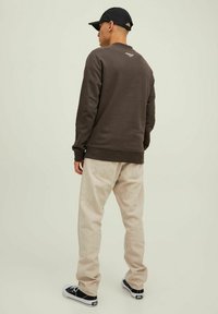 Brown sweatshirt with ribbed cuffs, beige jeans, and black sneakers. The sweatshirt features a small logo on the back. Casual, relaxed fit.
