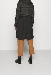 Chaqueta negra acolchada con costuras horizontales, superpuesta sobre un impermeable negro. Pantalones marrones recortados y botines negros completan el conjunto.