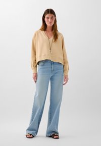 Beige Cropped-Sweatshirt mit V-Ausschnitt und langen Ärmeln, kombiniert mit hellblauen weiten Jeans. Schwarze Sandalen mit goldenen Akzenten.