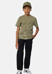 T-shirt verde oliva con motivo a macchie e logo nero, abbinato a pantaloni neri e sneakers bianche, completato con un cappellino coordinato.