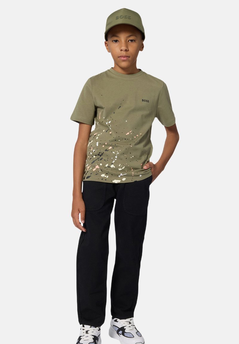 T-shirt verde oliva con motivo a macchie e logo nero, abbinato a pantaloni neri e sneakers bianche, completato con un cappellino coordinato.