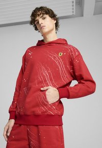 Sweatshirt rouge avec capuche, présentant un motif de taches blanches et un petit logo brodé. Fabriqué en tissu texturé, avec une poche kangourou.