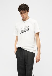 Νέος άνδρας που φοράει λευκό μπλουζάκι με σχέδιο αυτοκινήτου, μαύρο παντελόνι φόρμας Puma και ασημένιο κολιέ αλυσίδα, όρθιος με τα χέρια στις τσέπες.