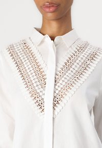 Camicia bianca con bottoni, caratterizzata da un dettagliato design in pizzo nella zona del petto, realizzata in tessuto fresco e con colletto strutturato.