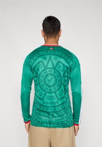 Man som bär en grön långärmad tröja med en detaljerad aztekisk kalenderdesign på ryggen och beige byxor, stående mot en vit bakgrund.