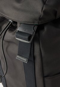 Gros plan d'une sangle de sac à dos noire avec une boucle en plastique gravée du logo « cK » sur un tissu foncé.