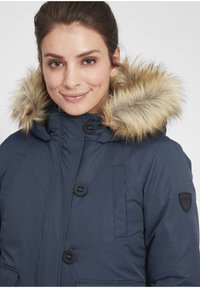 Tamsiai mėlynas izoliuotas parka su dirbtinio kailio apdaila ant gobtuvo, grioveliais tekstūra, dideliais priekiniais mygtukais ir šoniniais kišenėmis. Ant rankovės yra logotipo užrašas.