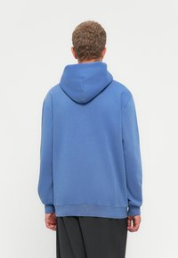 Hombre con cabello rizado corto, vistiendo una sudadera azul holgada y pantalones oscuros, de pie frente a un fondo blanco liso.