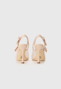 Sandales à talons hauts beiges avec une sangle en tissu texturé, un nœud décoratif et des accents de rhinestones transparents sur un panneau latéral transparent.