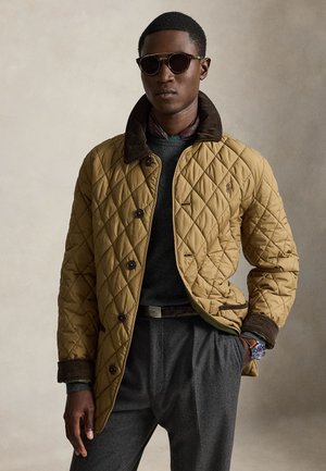 Mann mit Sonnenbrille, gesteppter hellbrauner Jacke mit dunklem Kragen, grauer Hose, dunklem Pullover, gemustertem Schal und Armbanduhr vor neutralem Hintergrund.