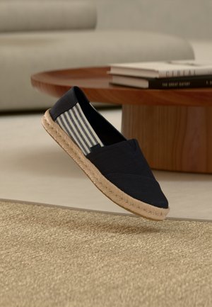 Chaussure bleu marine à enfiler avec doublure intérieure rayée et semelle tissée flottant au-dessus d'un tapis beige texturé dans un salon moderne.