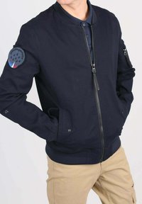Homme portant une veste bomber bleu marine avec des poches zippées et un patch de la Patrouille de France, associée à un pantalon beige.