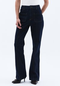 Tummansinisiä farkkuhousuja, joissa on kaksi etutaskua, näkyvät ompeleet, korkea vyötärö ja hyvin istuva malli nilkkoihin saakka.