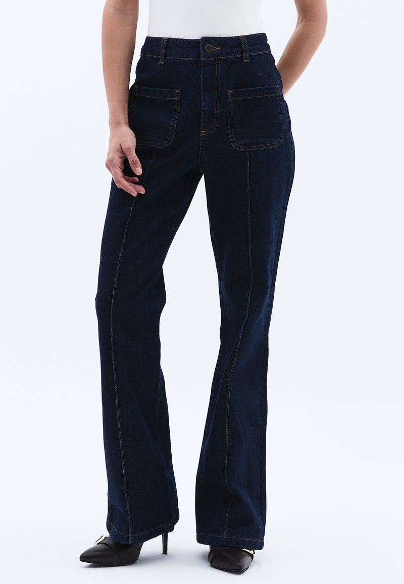 Tummansinisiä farkkuhousuja, joissa on kaksi etutaskua, näkyvät ompeleet, korkea vyötärö ja hyvin istuva malli nilkkoihin saakka.