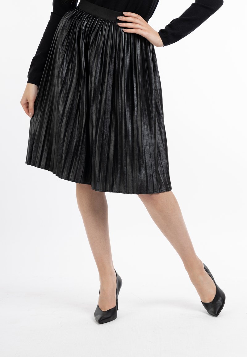 Sort plisseret nederdel lavet af glinsende materiale, med en bred talje. Båret med sorte højhælede sko, viser stoffet en glat overflade.