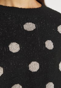 Svart stickad tröja med stora silverfärgade prickar, med en strukturerad yta och ribbad halsringning. Diskret metalltråd genom hela.
