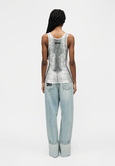 Γυναίκα με μακριά πλεξούδα, που φοράει ασημί tank top Moschino με υφή και ανοιχτόχρωμο τζιν παντελόνι φαρδιάς γραμμής, στέκεται με την πλάτη γυρισμένη.