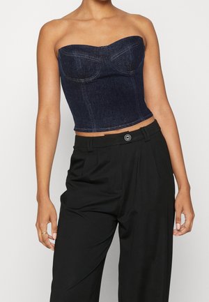 Kobieta w ciemnym denimu bez ramiączek, w górnej części bustier, zestawiona z wysokim stanem czarnych, szerokich spodni na tle jednolitego tła.