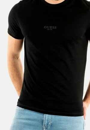 Homme portant un t-shirt noir ajusté Guess avec logo et un jean bleu clair, les mains partiellement dans les poches, sur un fond clair uni.