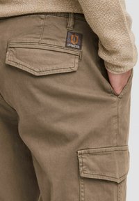 Beige cargo pants featuring a textured fabric, back pockets, a brand tag, and a side cargo pocket for additional storage.