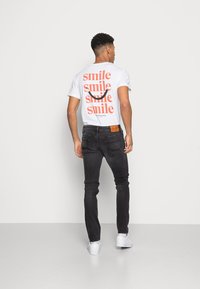 T-shirt branca com design de texto "sorriso" em vermelho, combinada com jeans pretos ajustados e tênis brancos. Ajuste casual, material em algodão.