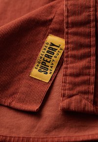 Tissu en velours côtelé couleur rouille avec des rainures verticales prononcées. Présente une étiquette jaune avec un texte noir indiquant "SUPERDRY HEAVY DUTY."