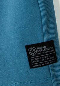 T-shirt de sport en teal avec une texture lisse, comportant une étiquette noire avec un texte blanc indiquant la marque et les instructions de lavage.