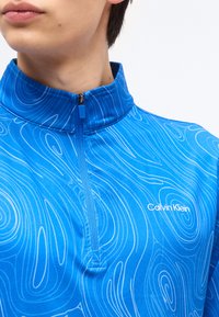 Νεαρό άτομο που φοράει μπλε επάνω μέρος Calvin Klein με φερμουάρ και λευκά αφηρημένα γραμμικά σχέδια σε ψηλό γιακά.