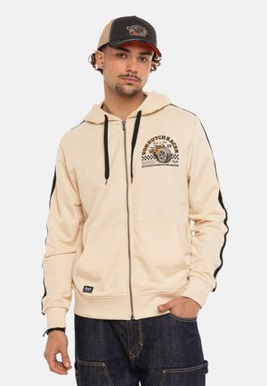 Jeune homme portant un sweat à capuche crème zippé avec le logo « Von Dutch Racer » et une casquette noir-beige, debout avec une main dans la poche.
