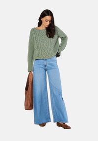 Grüner Strickpullover mit Zopfmuster, kombiniert mit hellblauen weit geschnittenen Jeans. Accessoires sind eine braune Tragetasche und braune Stiefeletten.