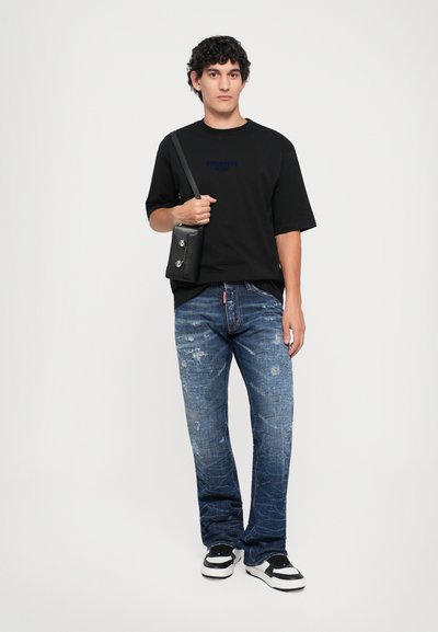 Tânăr îmbrăcat cu un tricou supradimensionat negru, blugi albaştri deterioraţi, adidaşi alb-negru şi ţinând o geantă neagră de umăr.