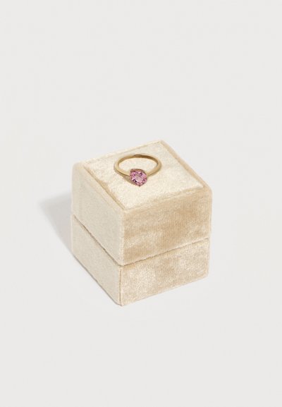 Χρυσό δαχτυλίδι με ροζ καρδιόσχημη πολύτιμη πέτρα, τοποθετημένο σε μπεζ βελούδινο κουτί κοσμημάτων πάνω σε λευκό φόντο.