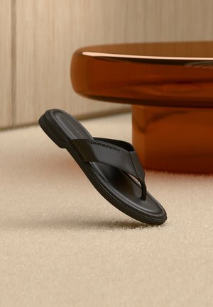 Schwarze Ledersandale mit Zehensteg von Tommy Hilfiger auf beigem Teppich, mit einem runden, bernsteinfarbenen modernen Tisch im Hintergrund.