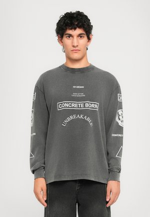 Young man with curly hair wearing a washed black sweatshirt with white text "CONCRETE BORN UNBREAKABLE" and graphic prints on sleeves.
