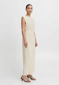 Pleated, pitkä beige mekko korkealla kaula-aukolla ja ilman hihoja. Ominaisuuksia on joustava vyötärö ja se ulottuu nilkkoihin. Yhdistetty mustiin hihnakiiltoihin.