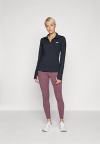 Maglietta sportiva nera a maniche lunghe con mezza zip, abbinata a leggings viola e sneaker bianche, su uno sfondo neutro.